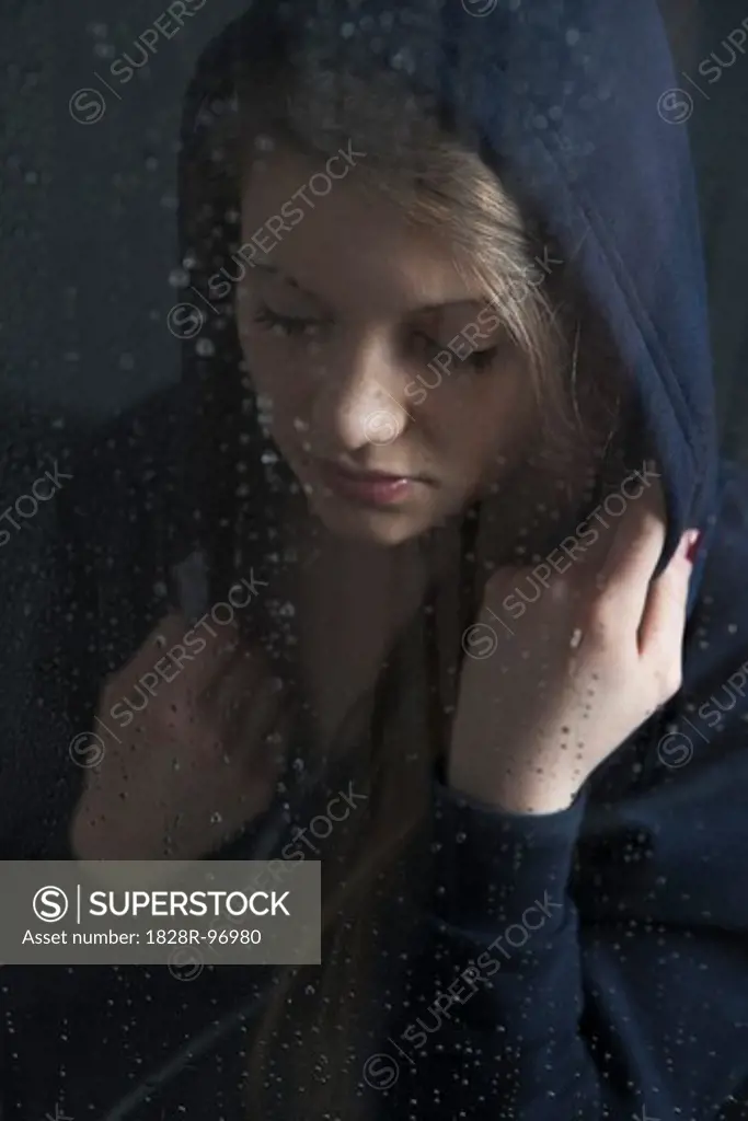 Portrait of young woman behind window, wet with raindrops, wearing hoodie, looking down,05/05/2013