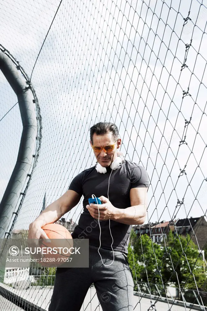 Mature man standing on outdoor basketball court holding basketball and looking at MP3 player, Germany,05/02/2013