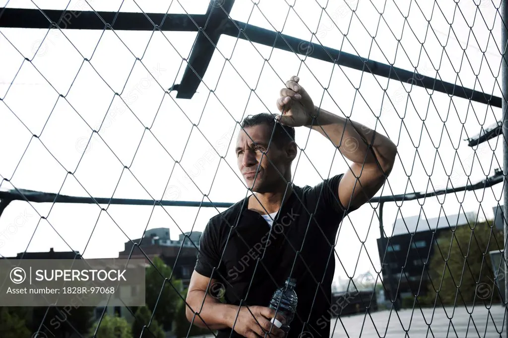 Mature man leaning against chain-link fence on outdoor basketball court, holding bottle of water, Germany,05/02/2013