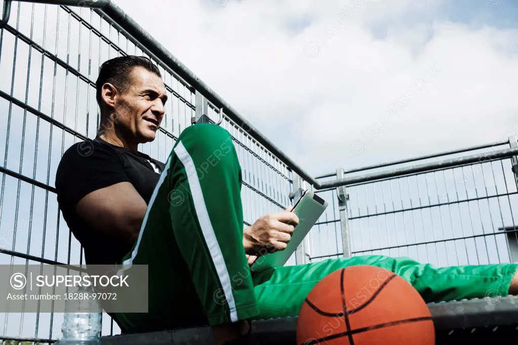Mature man sitting at top of stairs on outdoor basketball court looking at tablet computer, Germany,05/02/2013