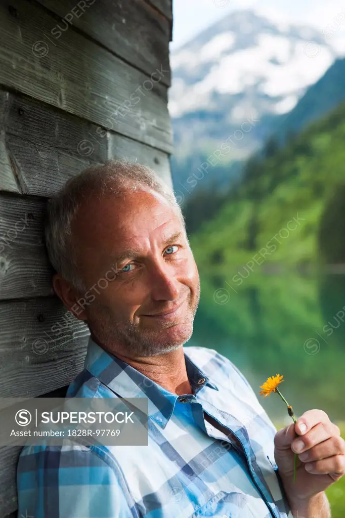Portrait of mature man looking at camera, holding flower, standing next to building at Lake Vilsalpsee, Tannheim Valley, Austria,06/15/2013