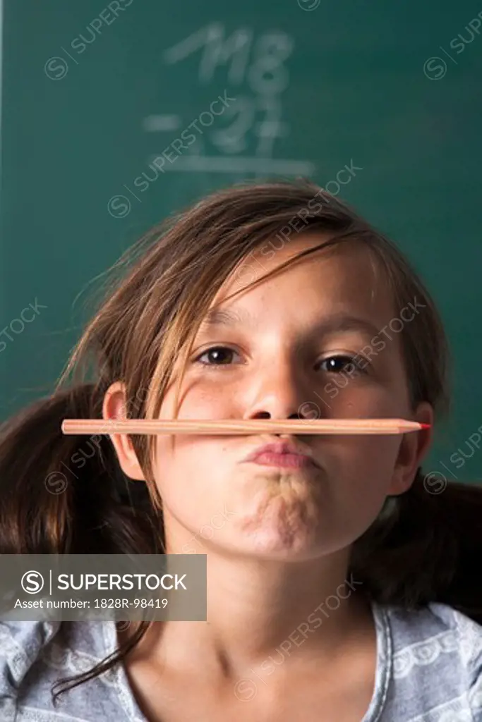 Portrait of girl standing in front of blackboard in classroom, holding pencil with mouth, Germany,07/24/2013