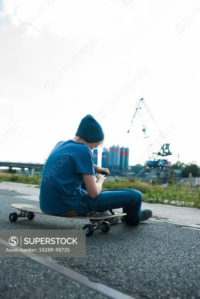Backview of boy sitting on skateboard outdoors in an industrial area and looking at cell phone, Germany,08/21/2013
