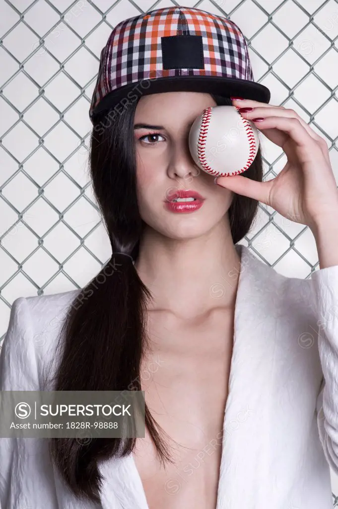 Close-up portrait of young woman wearing baseball cap and holding baseball in front of eye, studio shot on white background,04/05/2013