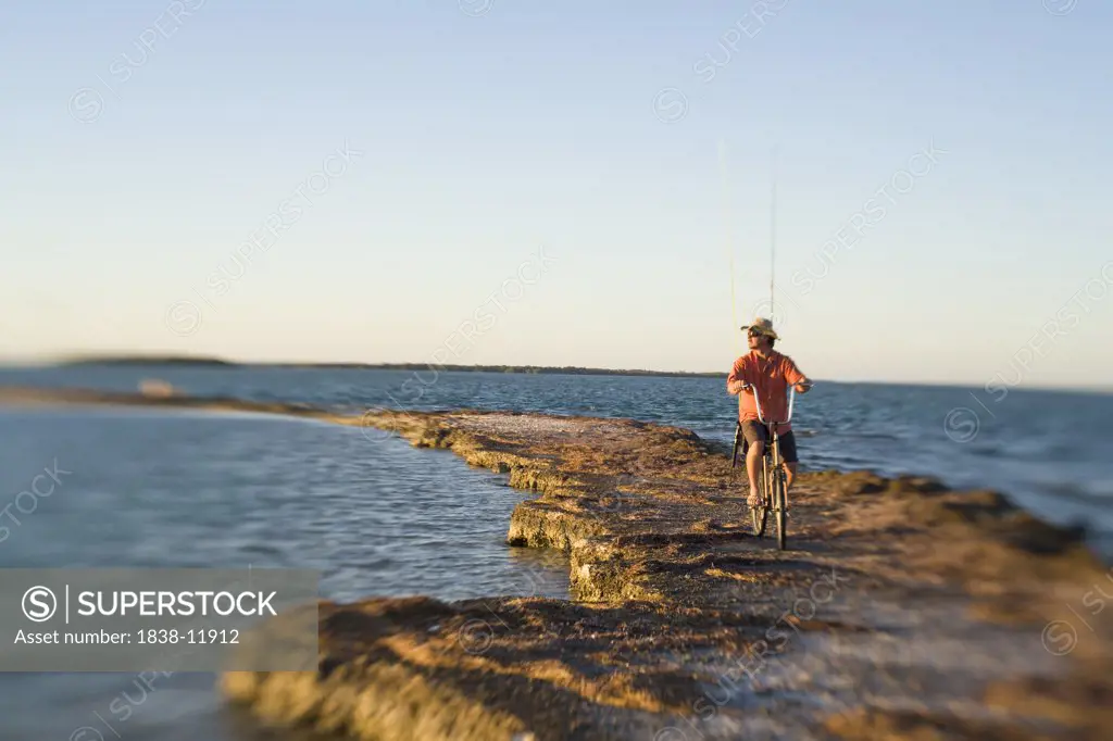 Man Riding Bicycle With Fishing Rods on Rocky Jetty, Florida Keys, USA
