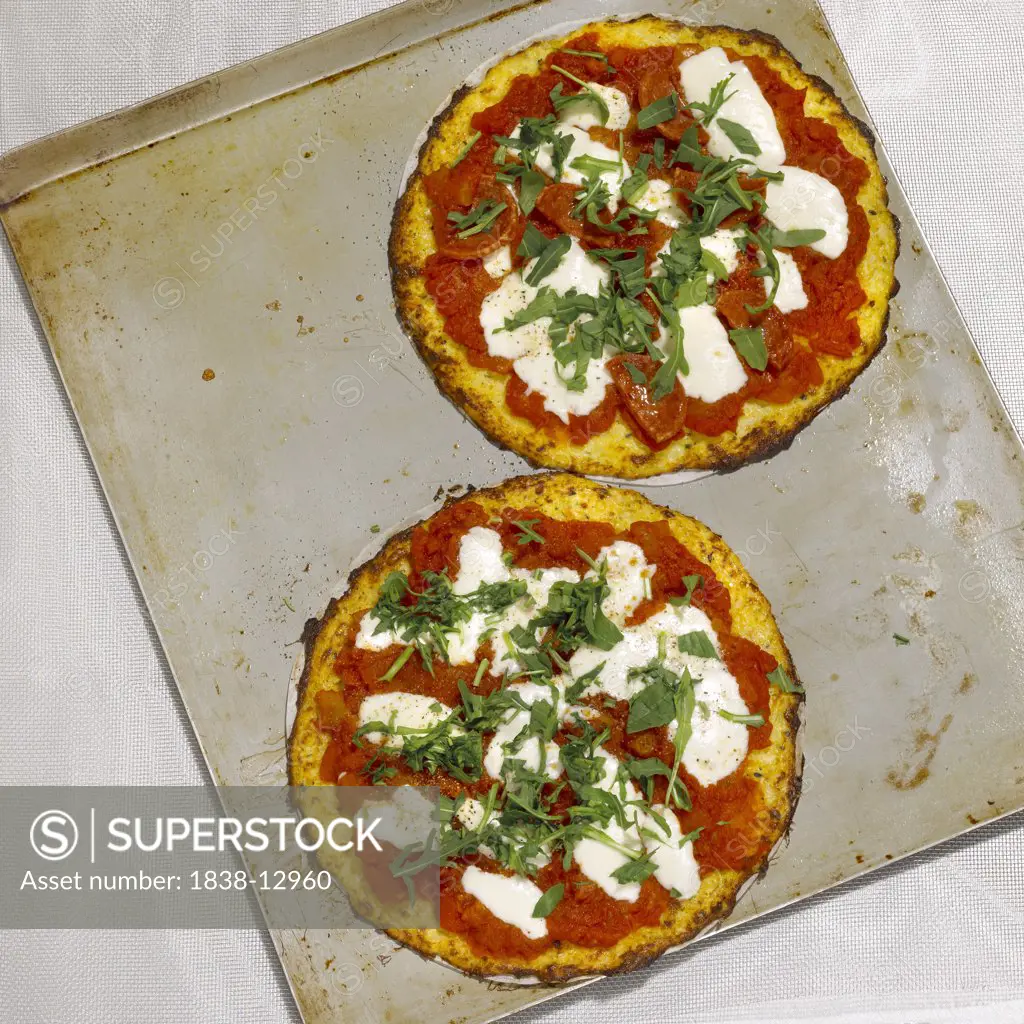 Two Small Cauliflower Crust Pizzas Topped with Arugula on Baking Sheet