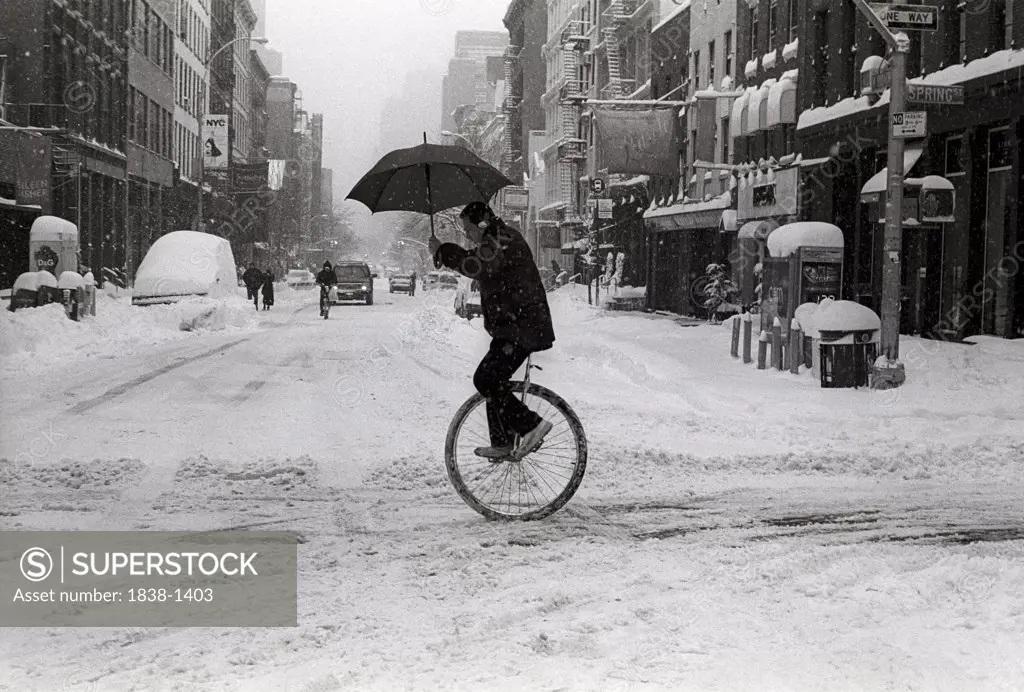Man riding a unicycle in the snow, holding an umbrella, in winter.