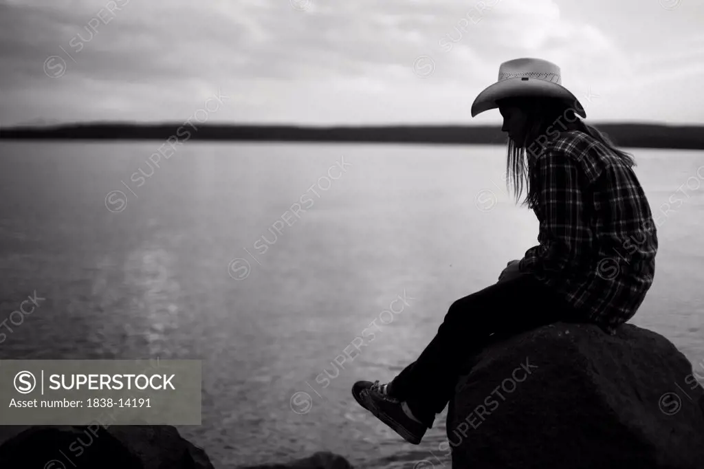 Young Woman in Cowboy hat Sitting on Rock Near Lake, Yellowstone National Park, USA