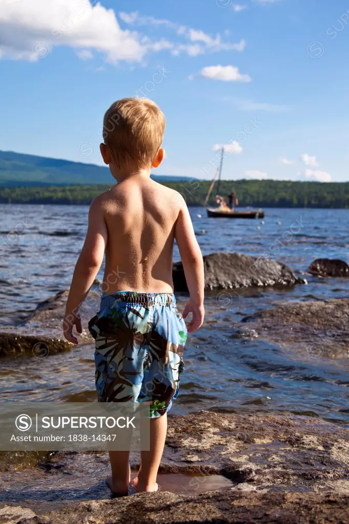 Young Boy in Bathing Suit Standing on Rocks at Water's Edge