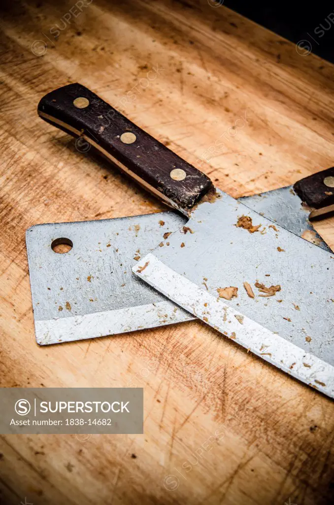 Two Meat Cleavers Covered with Bits of Meat on Wooden Cutting Board