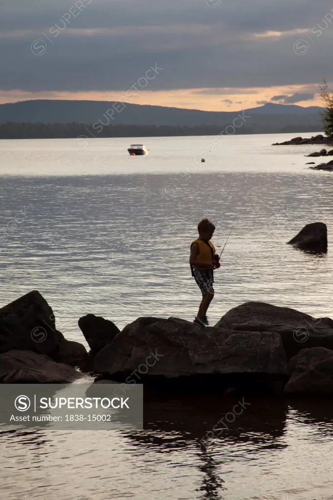 Young Boy Fishing off Rocks in Lake at Sunset