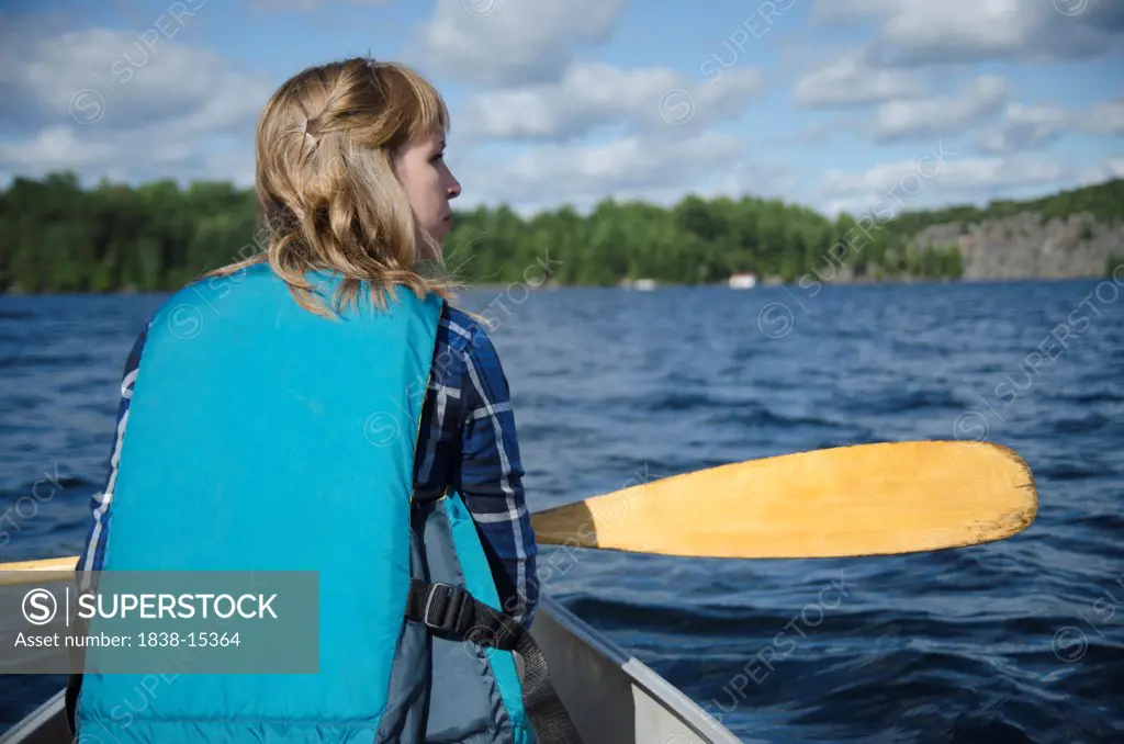 Young Woman in Life Jacket Holding Paddle and Sitting in Canoe on Lake Looking Out