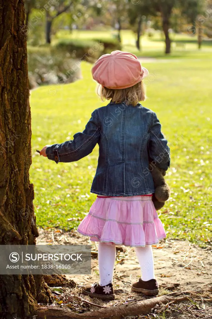 Young Girl Holding Stuffed Animal Standing Next to Large Tree in Park, Rear View