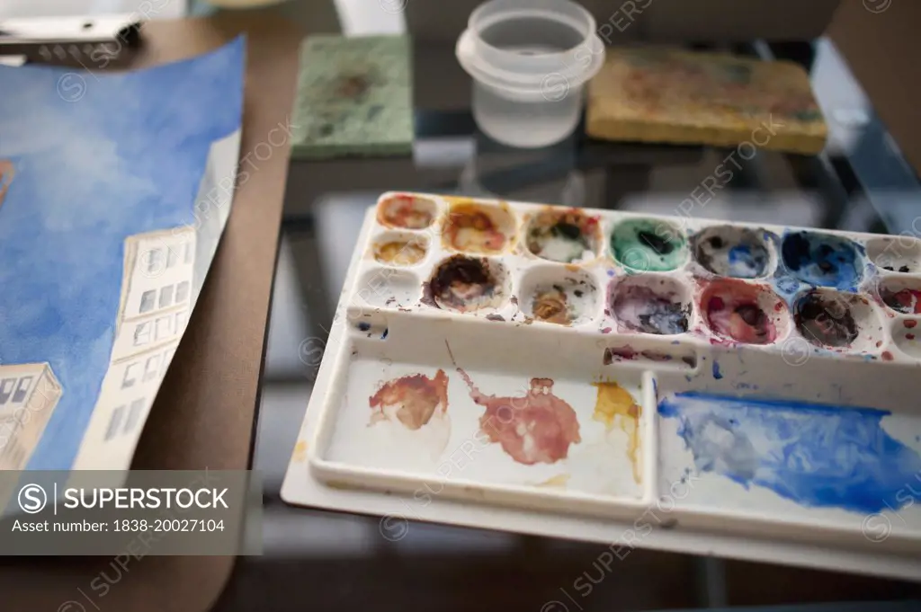 Messy Watercolor Palette Next to Watercolor Painting on Artist's Desk, Close-Up