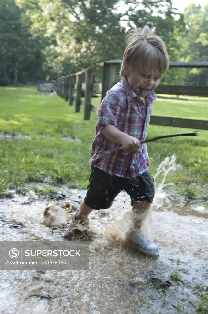 Smiling Young Boy Running Through Puddle and Carrying Stick