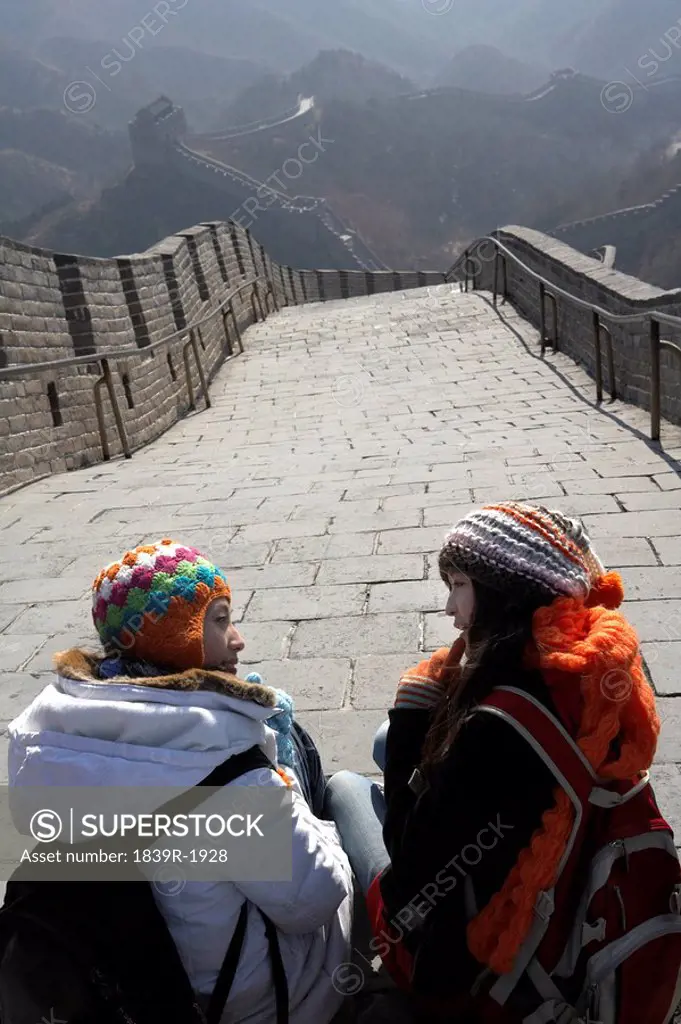 Young Women Sitting On The Great Wall Of China Smiling At Each Other