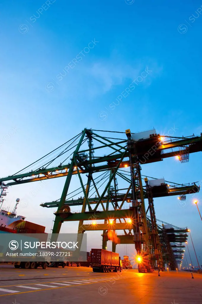 Cranes, cargo containers and trucks at a shipping port during dusk