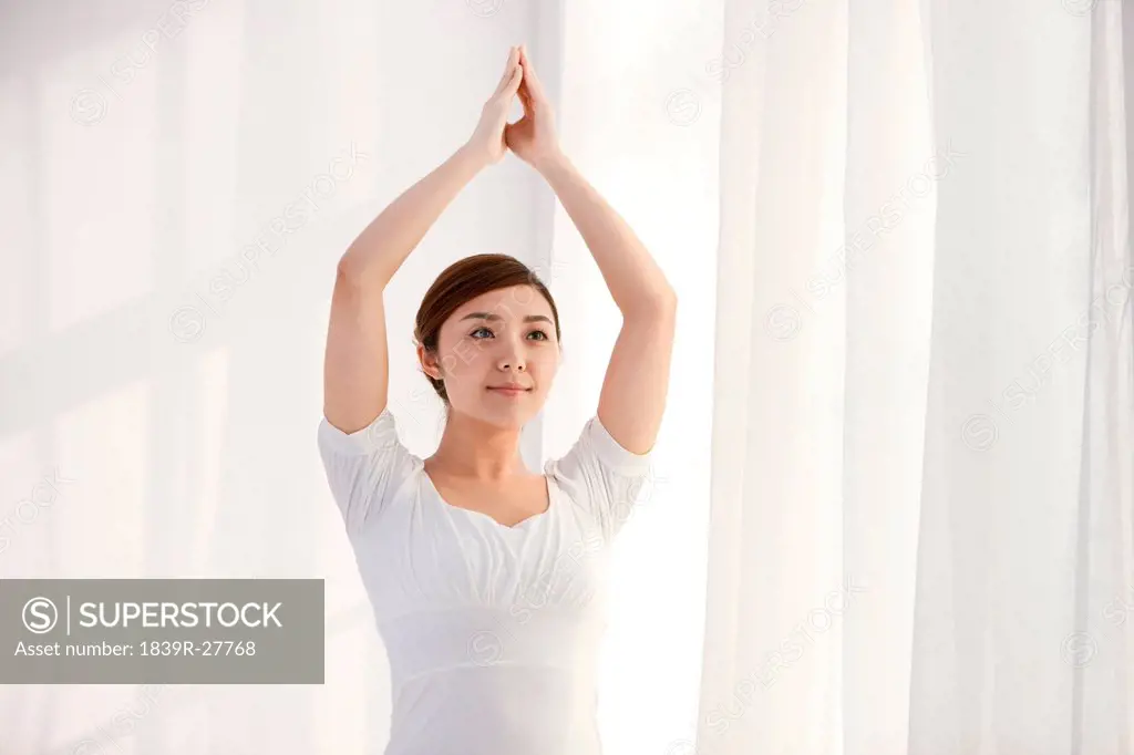 Young woman practicing yoga by a window with arms raised
