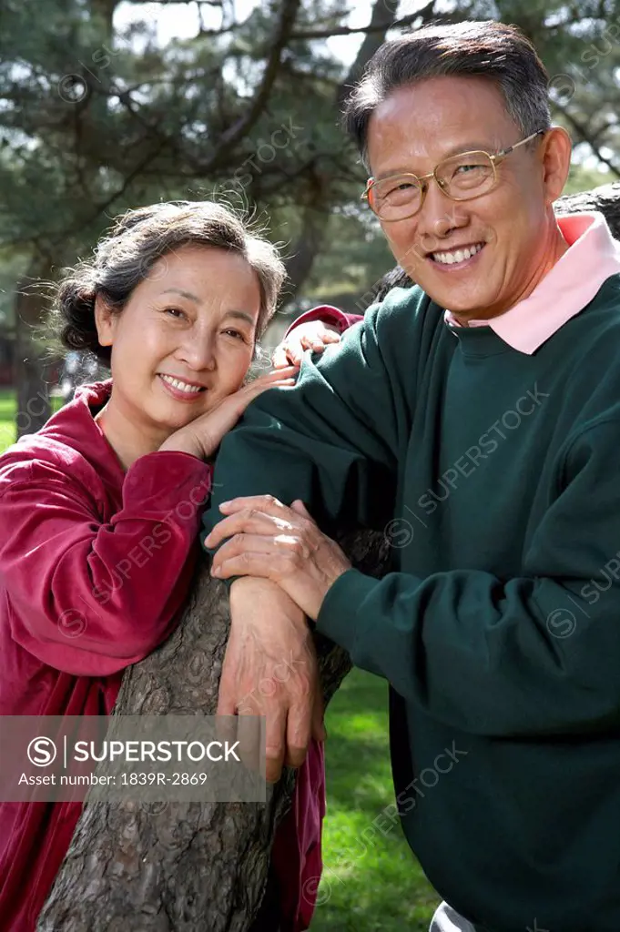 Chinese Couple Leaning On Tree Branch In Park Both Looking And Smiling At The Camera