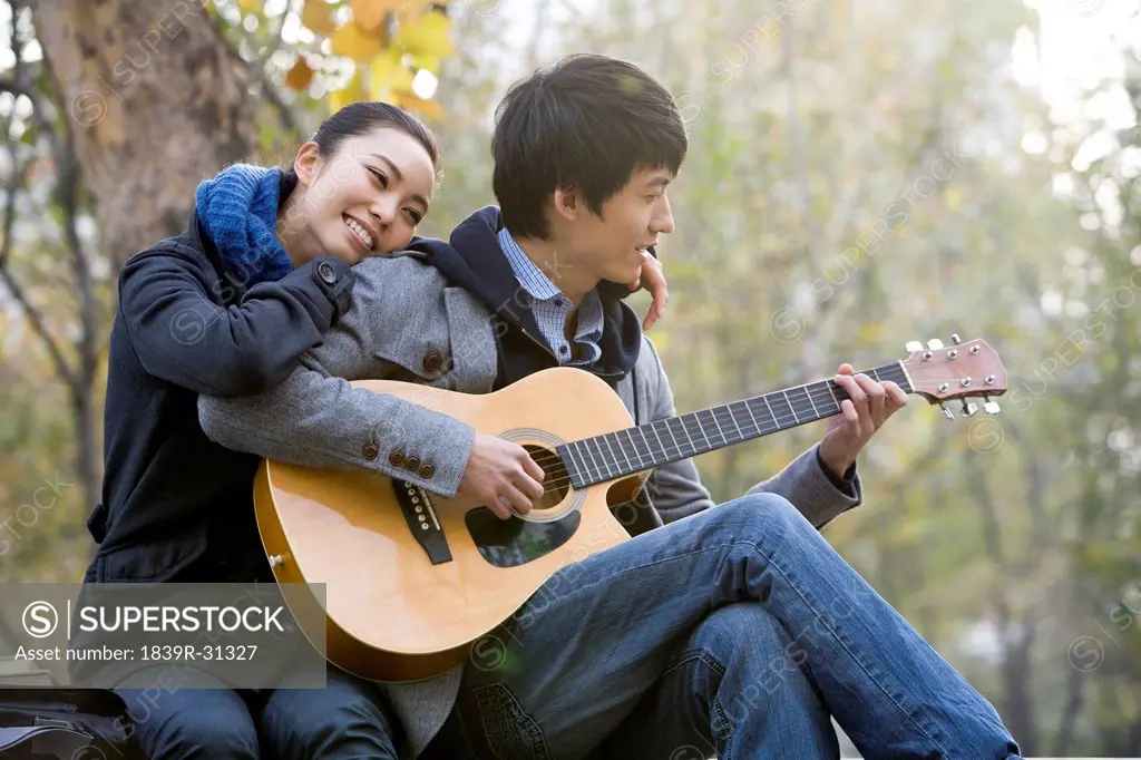 A young man plays the guitar as he girlfriend leans on him affectionately