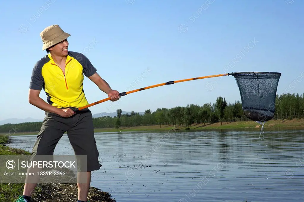 Man Standing On A Lake Bank Having Caught A Fish In His Net