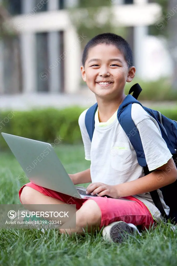 Smiling schoolboy sitting on the lawn with laptop
