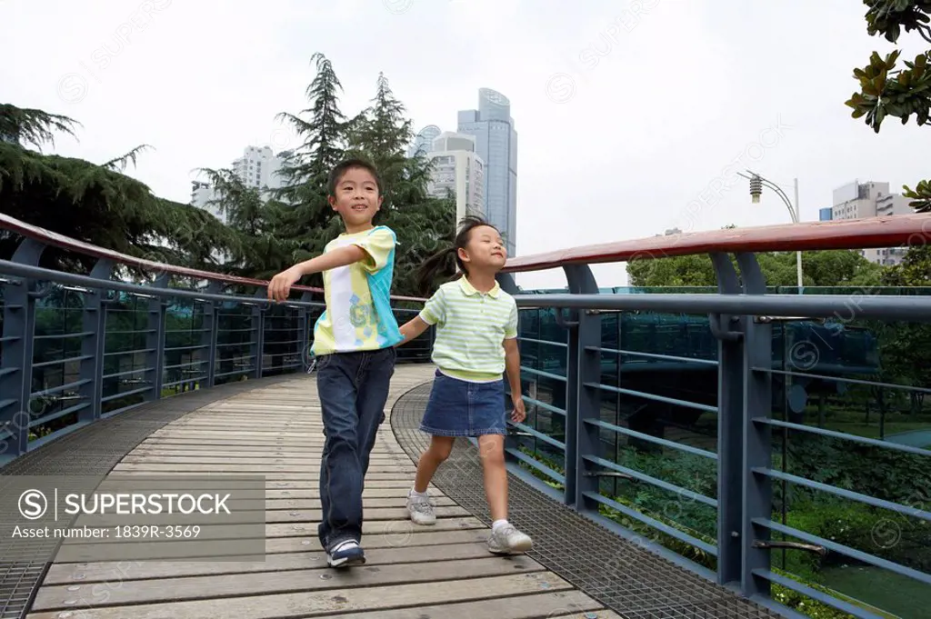 Young Boy And Young Girl Running Across A Bridge In A Park
