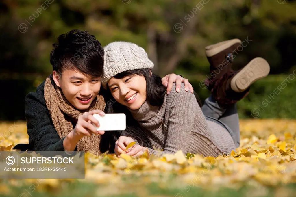 Sweet young couple with smart phone lying on golden autumn leaves