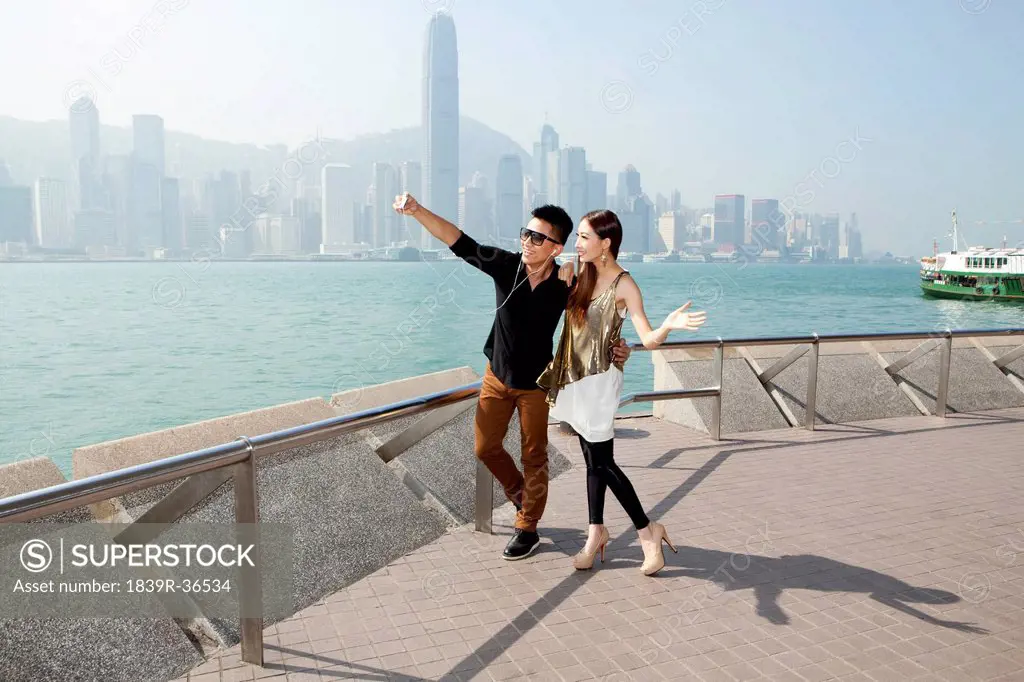 Fashionable young couple taking self-portrait pictures with smart phone in Victoria Harbor, Hong Kong