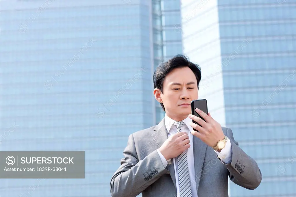 Serious businessman adjusting tie by reflection of mobile phone screen