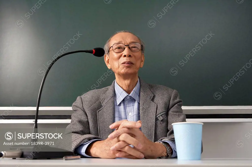 Teacher Sitting At Desk And Talking Into A Microphone