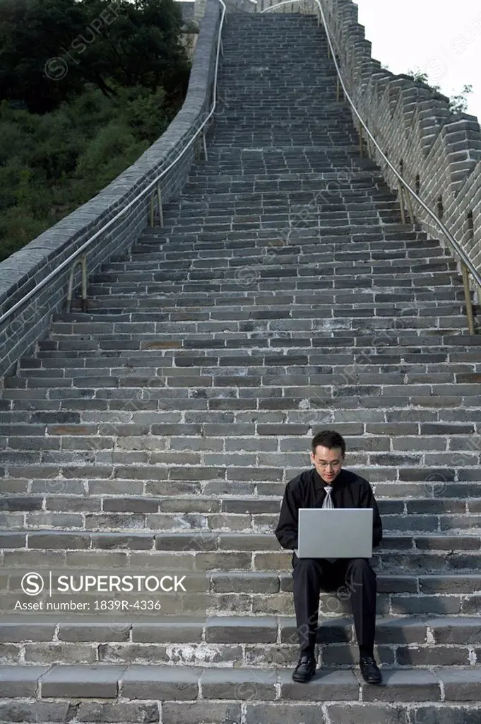 Businessman Sitting On Steps Of Great Wall Of China, Typing On Laptop Computer