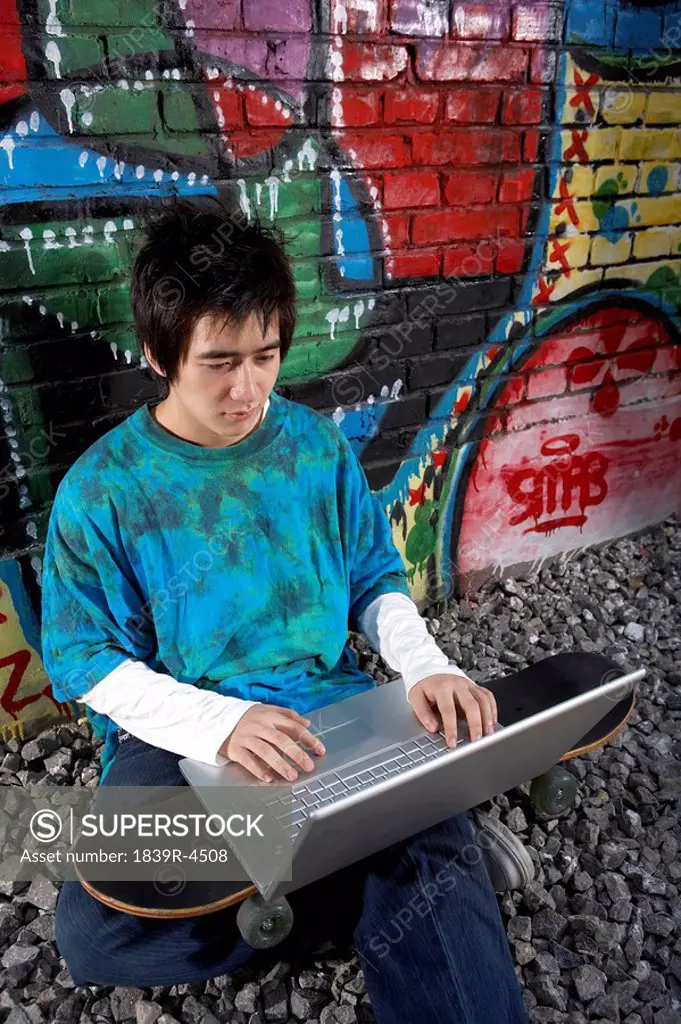 Teenage Boy Sitting With Skateboard And Laptop In Front Of A Wall Of Graffiti