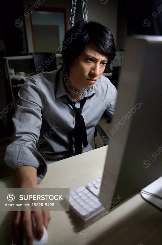 Young man in shirt and tie is hung from a chain at his cubicle while he uses his computer