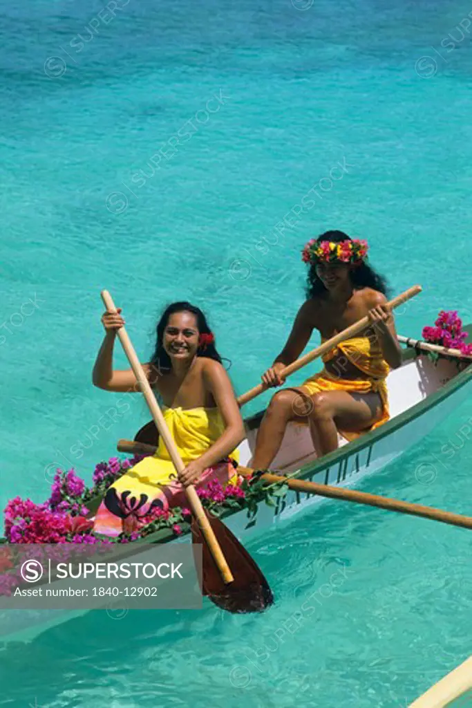Two young women in traditional clothing paddle an outrigger canoe across a turquoise lagoon on the island of Huahine in the Society Islands of French Polynesia.