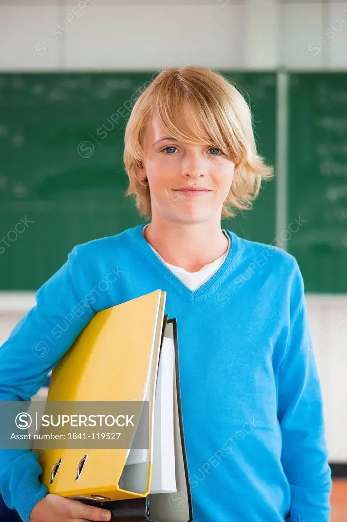 Smiling schoolboy in classroom holding file folders, portrait