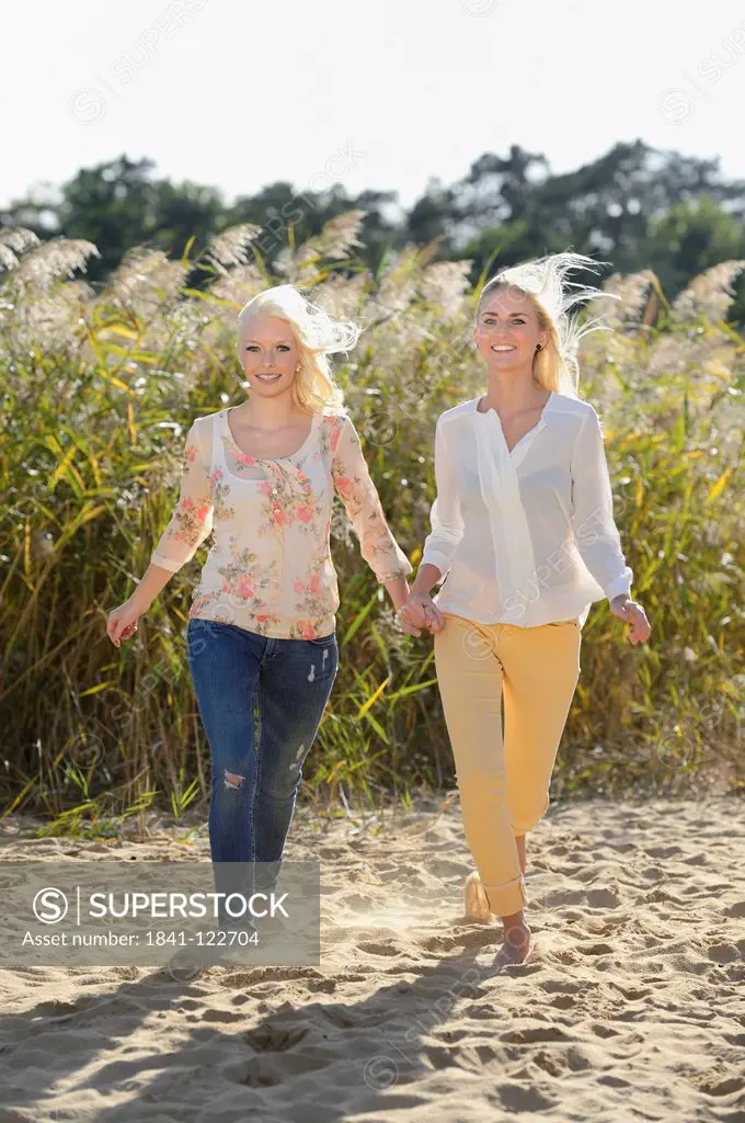 Two happy young blond women walking hand in hand on sandy beach