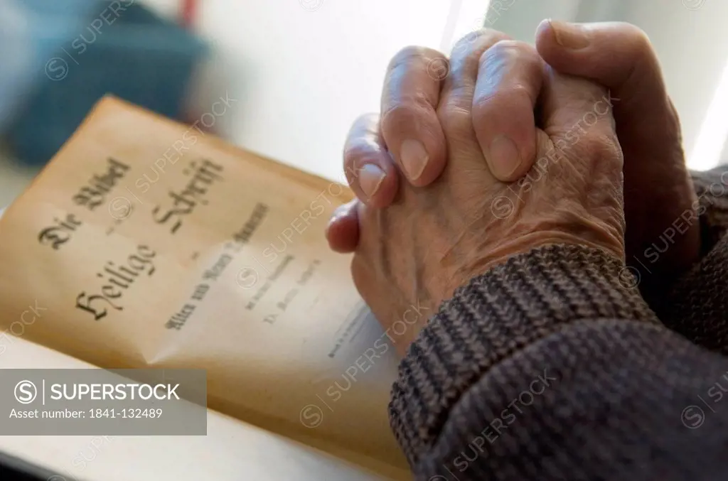 Praying hands of an elderly person held over the first page of an old bible.