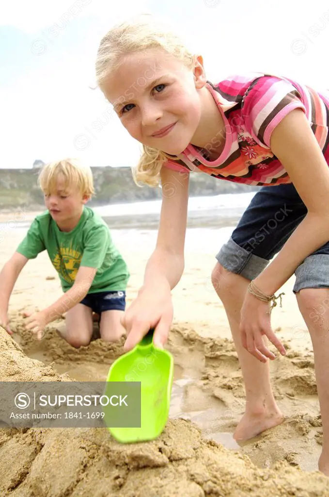 Portrait of girl holding shovel with her friend making sandcastle on beach