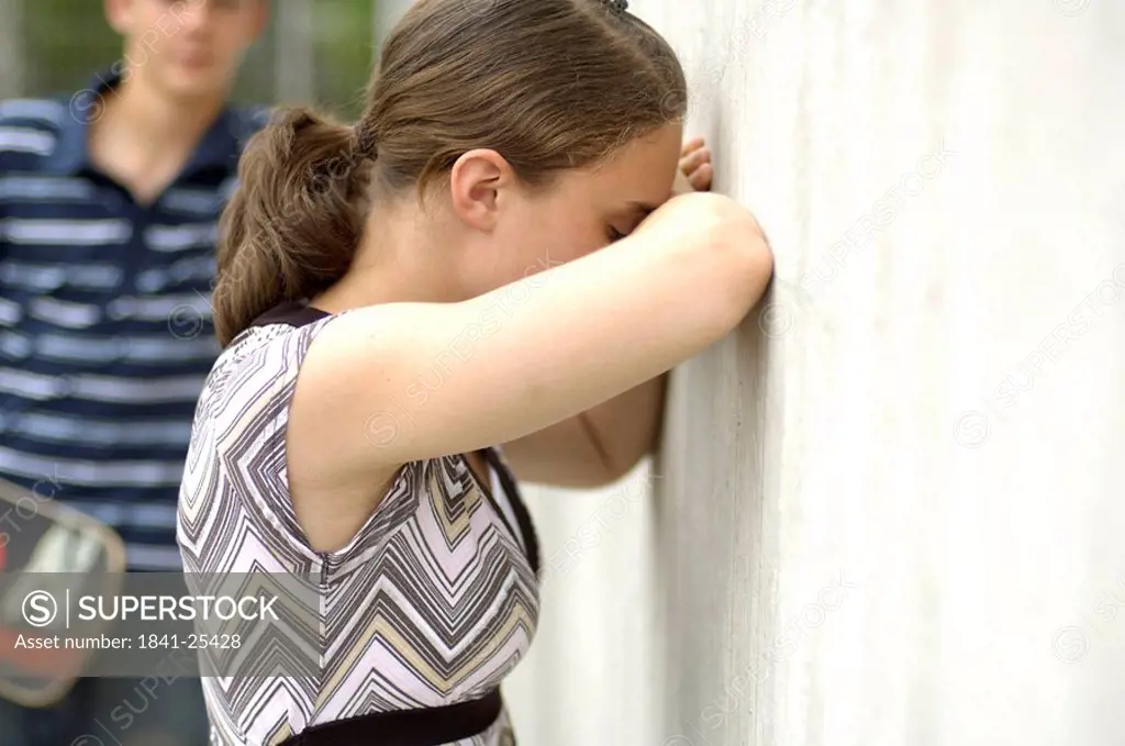 Side profile of teenage girl sobbing against wall with her friend in background