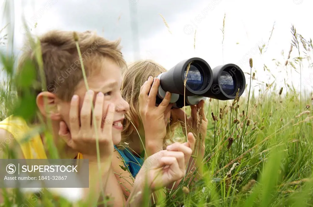 Boy looking through binoculars in field with his friend sitting beside him