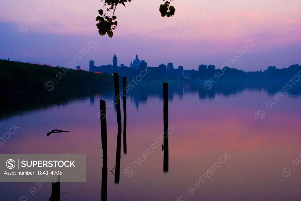 Wooden posts in lake with buildings in background at sunset, Lago Di Mezzo, Mantua, Lombardy, Italy
