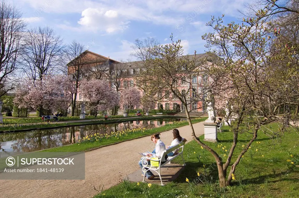 Two women sitting on bench near pool in front of palace, Kurfuerstliches Palais, Trier, Germany