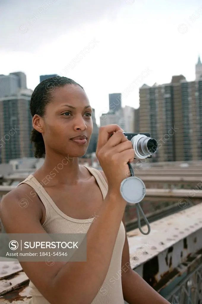 Woman standing on the Brooklyn Bridge and holding a digital camera, skyline of New York in the background, USA