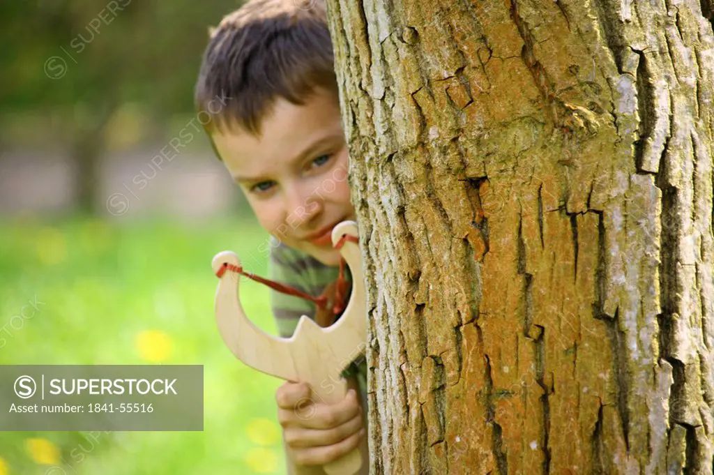 Close_up of boy hiding behind tree and using slingshot