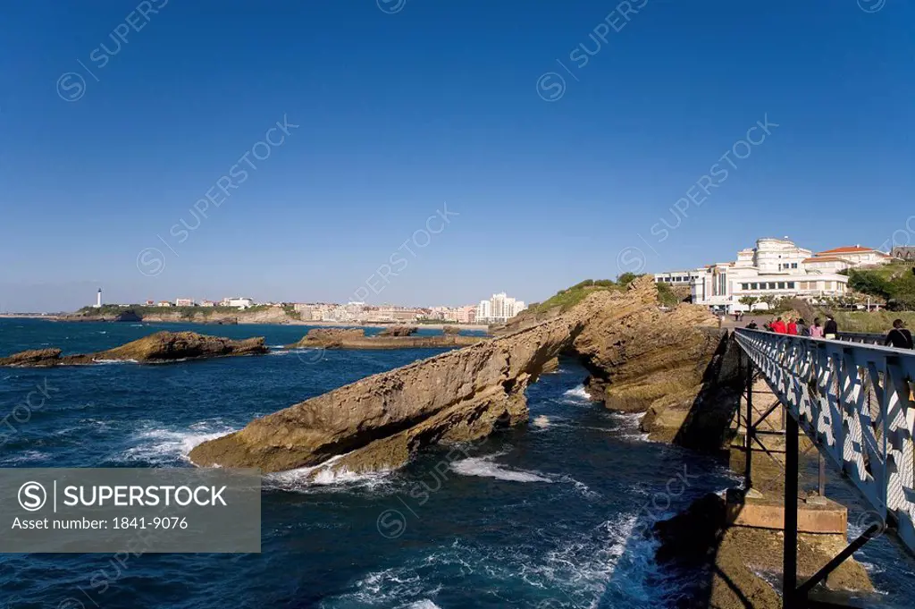 Tourists walking on bridge near natural arch with lighthouse in background, Pointe Saint_Martin, Biarritz, Pyrenees_Atlantiques, Aquitaine, France