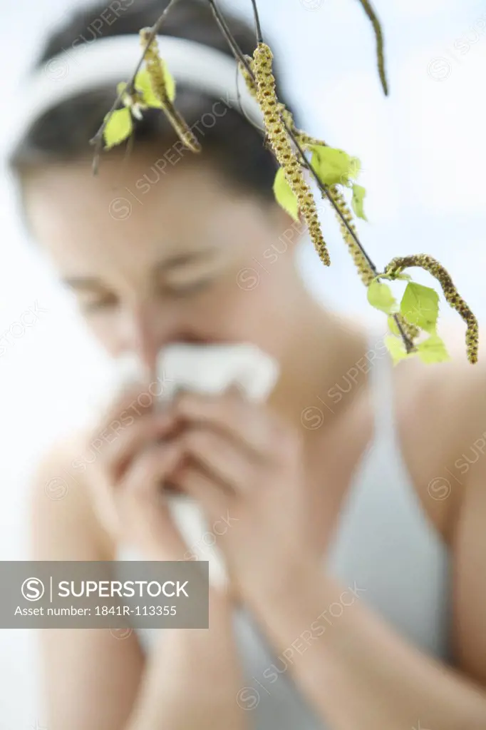 a branch with leaves of a birch tree - in the background blurred a young woman  sneezing and giving her nose a blow