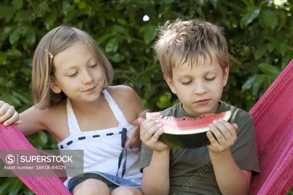 Boy and girl sitting in hammock, boy eating melon