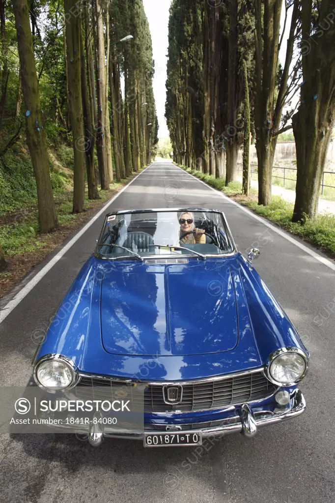 Senior woman driving convertible on tree-lined road, Italy
