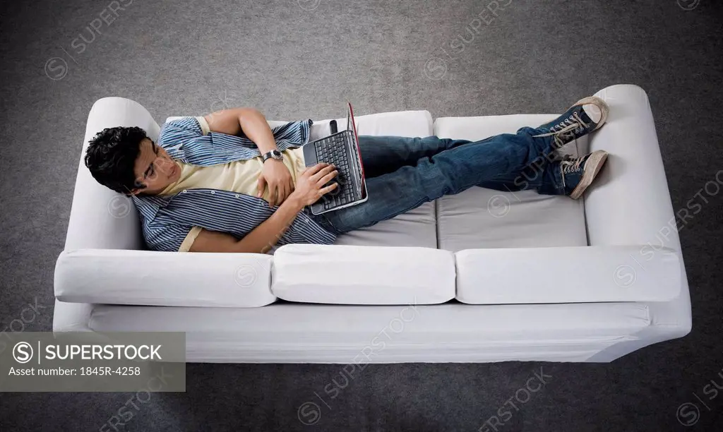 High angle view of a man lying on a couch working on a laptop